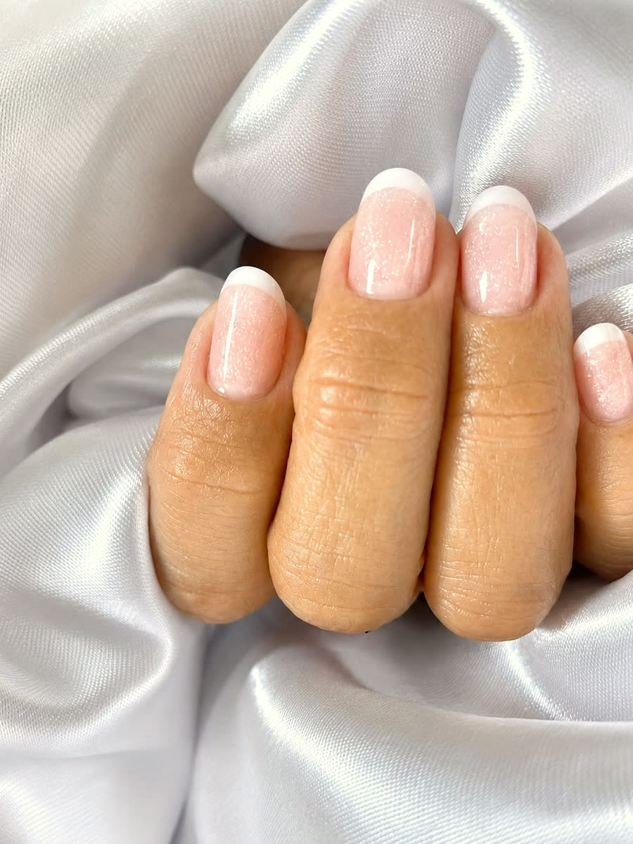 Unhas curtas com banho em gel nude e ponta branca, apoiadas em cetim branco